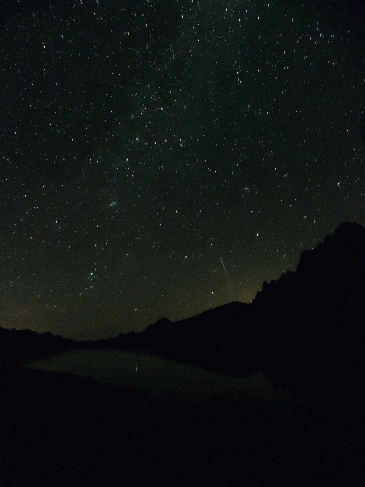 Photo de nuit du ciel au-dessus d'un lac ; on voit la crête qui se découpe sur un ciel étoilé et la trace d'une étoile filante.