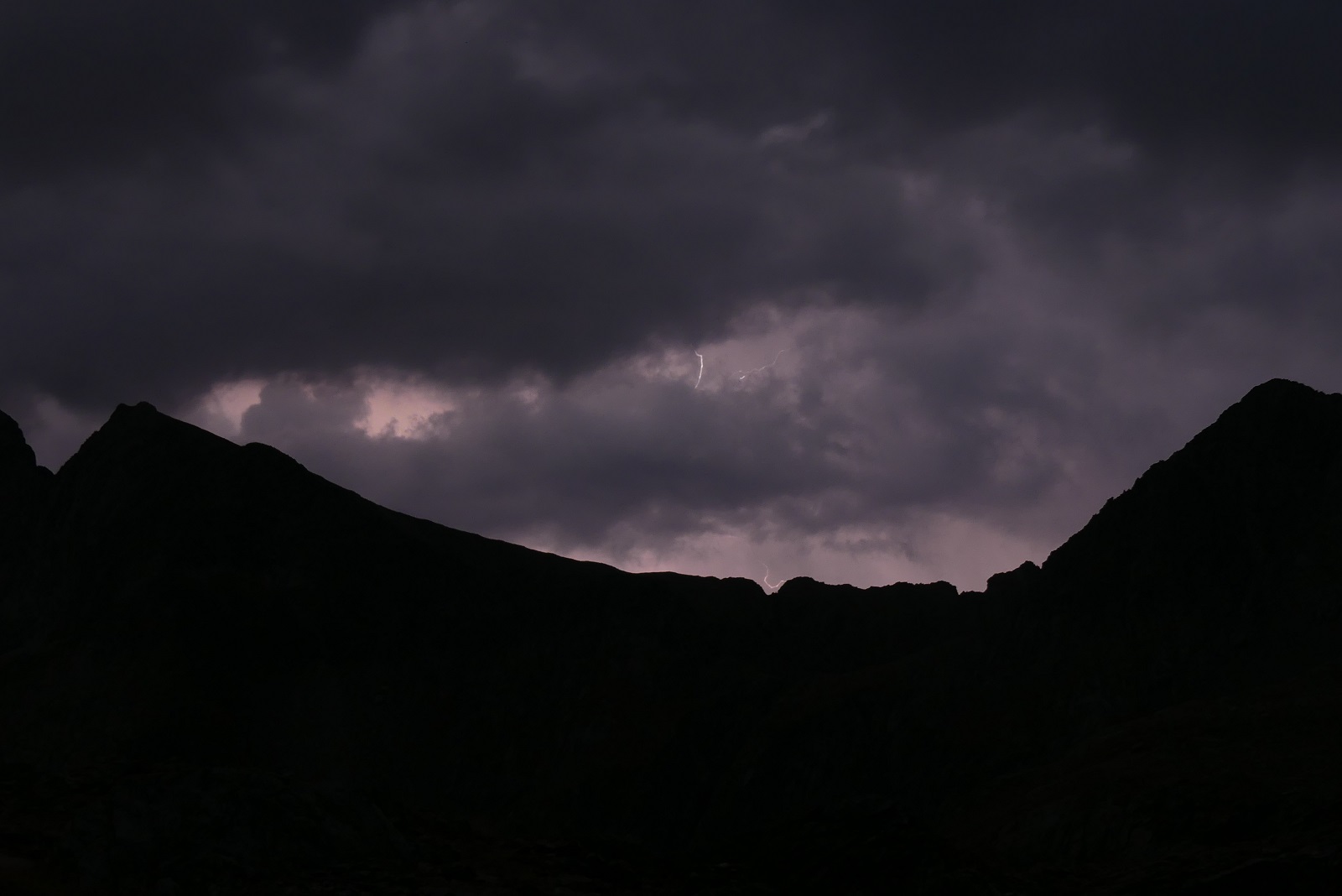 Photo du ciel de nuit, on voit le profil de la montagne devant un ciel violet foncé chargé de nuages. Un éclair apparaît entre deux nuages.